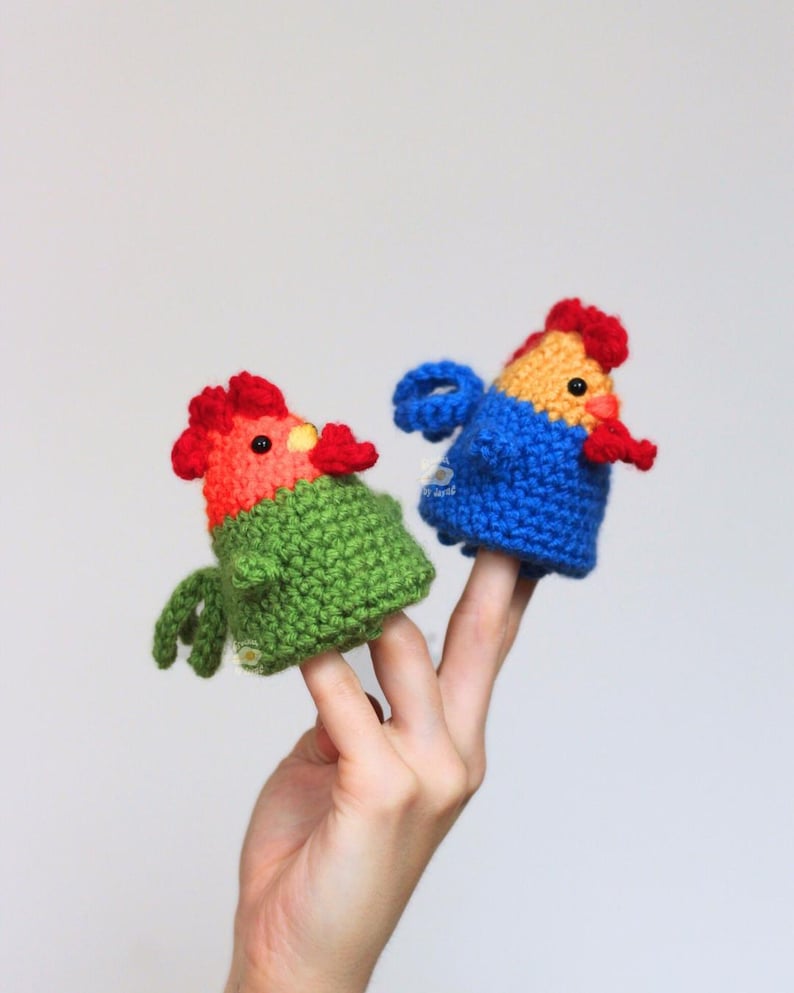 May include: Two handmade crochet finger puppets shaped like colorful roosters. One is green and red, the other is blue and yellow. The puppets are held up by a hand against a white background.