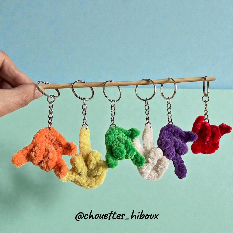 May include: A collection of six colorful crochet hand keychains, each with a different color: orange, yellow, green, white, purple, and red. The keychains are hanging from a bamboo stick with silver key rings and chains. The background is a light blue.
