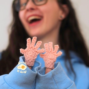 May include: Two small, peach-colored crochet hand puppets. The puppets are on fingers, with the text "Crochet by Jayne" on the blue sweatshirt. The puppets are made of a textured yarn and have detailed fingers.