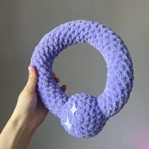 May include: A purple crocheted ring with a fluffy texture and two white stars on the side. The ring is held in a hand.