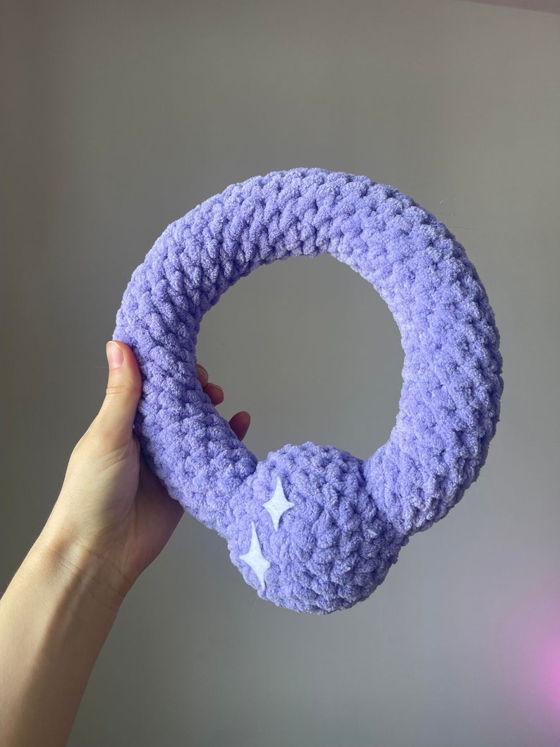 May include: A purple crocheted ring with a fluffy texture and two white stars on the side. The ring is held in a hand.