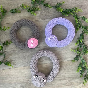 May include: Three crocheted rings with different colored fabric and embellishments. The first ring is gray with a pink flower, the second is purple with white stars, and the third is gray with a gray face.