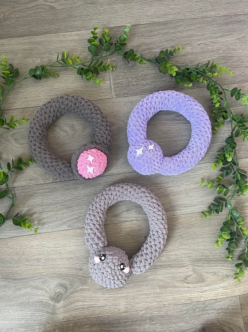 May include: Three crocheted rings with different colored fabric and embellishments. The first ring is gray with a pink flower, the second is purple with white stars, and the third is gray with a gray face.