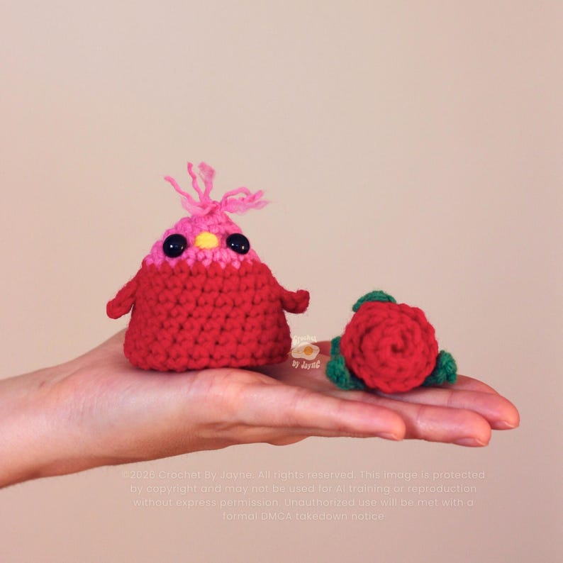Puede incluir: Un p&aacute;jaro de ganchillo hecho a mano, rojo y rosa, con ojos negros y un mech&oacute;n rosa, y una rosa de ganchillo roja con hojas verdes. El p&aacute;jaro tiene brazos peque&ntilde;os. Los art&iacute;culos se muestran en una mano.