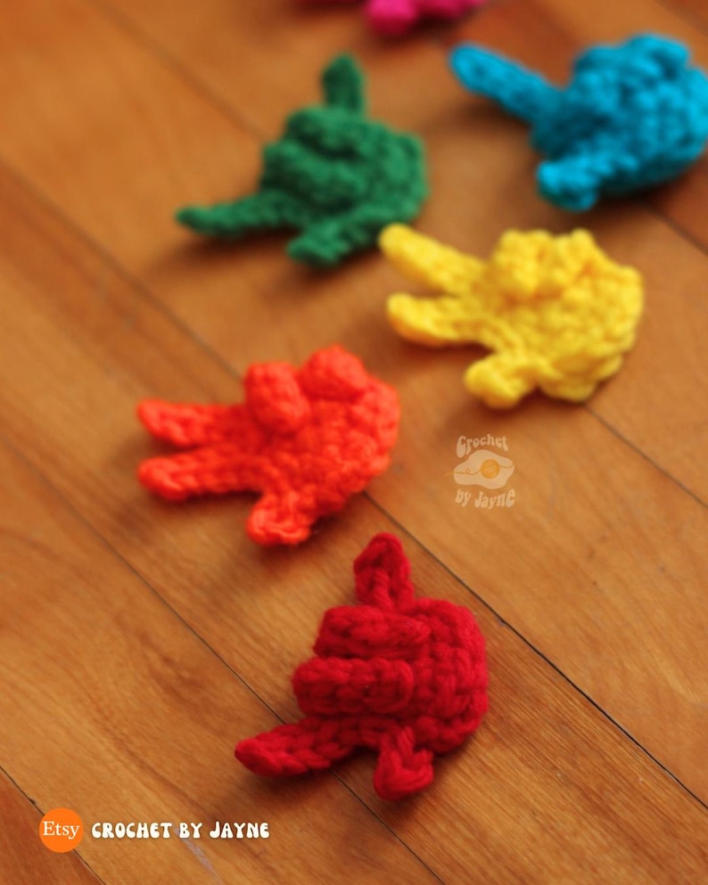 May include: A collection of colorful, crocheted hand-shaped items in red, yellow, green, and blue. The items are scattered on a wooden surface. The Etsy shop name "Crochet by Jayne" is visible in the image.