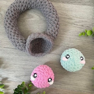 May include: Two small crocheted plush toys with embroidered eyes and pink and blue faces. A gray crocheted ring with a small gray crocheted bowl.