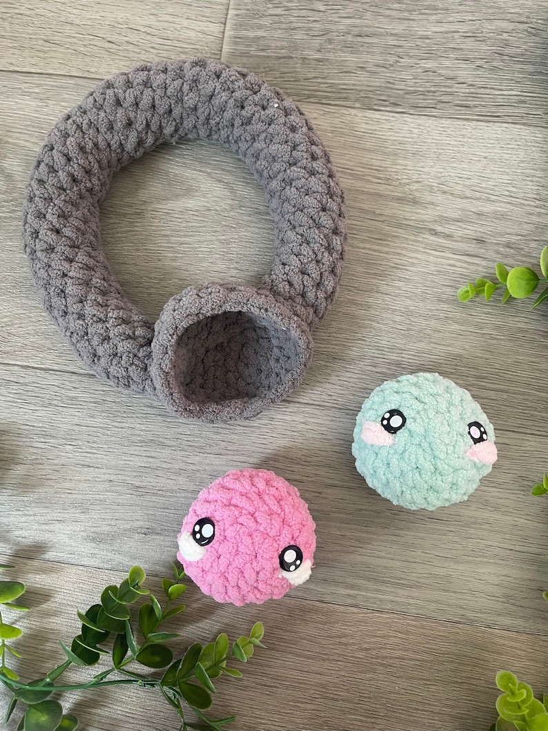 May include: Two small crocheted plush toys with embroidered eyes and pink and blue faces. A gray crocheted ring with a small gray crocheted bowl.