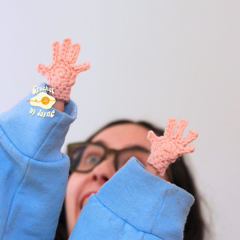 May include: Two small, peach-colored crochet hand puppets are displayed, one on each finger. The hands are detailed with individual fingers. The puppets are on a blue sleeve. The text "Crochet by Jayne" is visible.