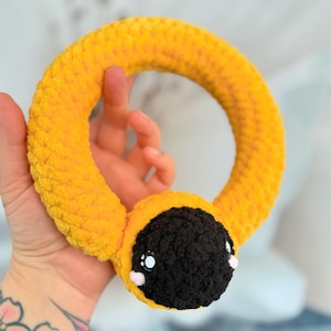 May include: A yellow crocheted ring toy with a black and white face. The toy has large eyes and a small pink nose.