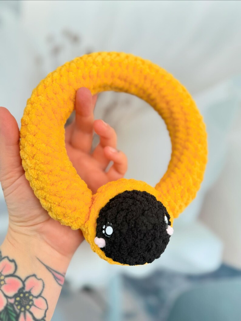 May include: A yellow crocheted ring toy with a black and white face. The toy has large eyes and a small pink nose.
