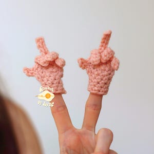 May include: Two pink crocheted finger puppets shaped like hands giving the middle finger. The puppets are worn on fingers, with the middle finger extended. The text "Crochet by Jayne" is visible on a small tag.