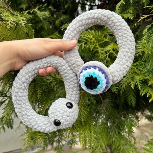 May include: A gray crocheted snake with a blue evil eye charm. The snake is curled into a circle and has two black eyes.