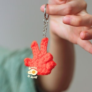 May include: A handmade, bright orange crochet keychain in the shape of a hand making the peace sign. The keychain is attached to a silver metal ring and chain. The text "Crochet by Jayne" is on the keychain.