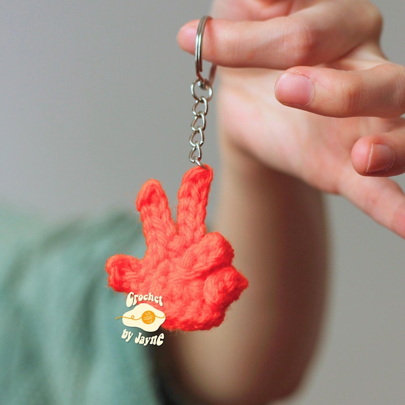 May include: A handmade, bright orange crochet keychain in the shape of a hand making the peace sign. The keychain is attached to a silver metal ring and chain. The text "Crochet by Jayne" is on the keychain.