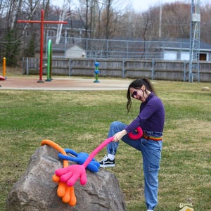 May include: A person wearing a purple shirt and blue jeans is pulling on a long, pink crocheted arm with a blue hand. The arm is attached to a large, gray rock. The crocheted arm is wrapped around the rock with a smaller orange crocheted arm and hand.