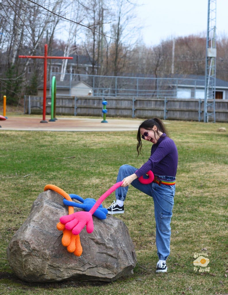 May include: A person wearing a purple shirt and blue jeans is pulling on a long, pink crocheted arm with a blue hand. The arm is attached to a large, gray rock. The crocheted arm is wrapped around the rock with a smaller orange crocheted arm and hand.