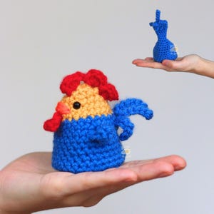 May include: A handmade crochet rooster in blue, yellow, and red, held in a hand. The rooster has a blue body, yellow head, and red comb and wattle. A smaller blue crochet figure is held in another hand.