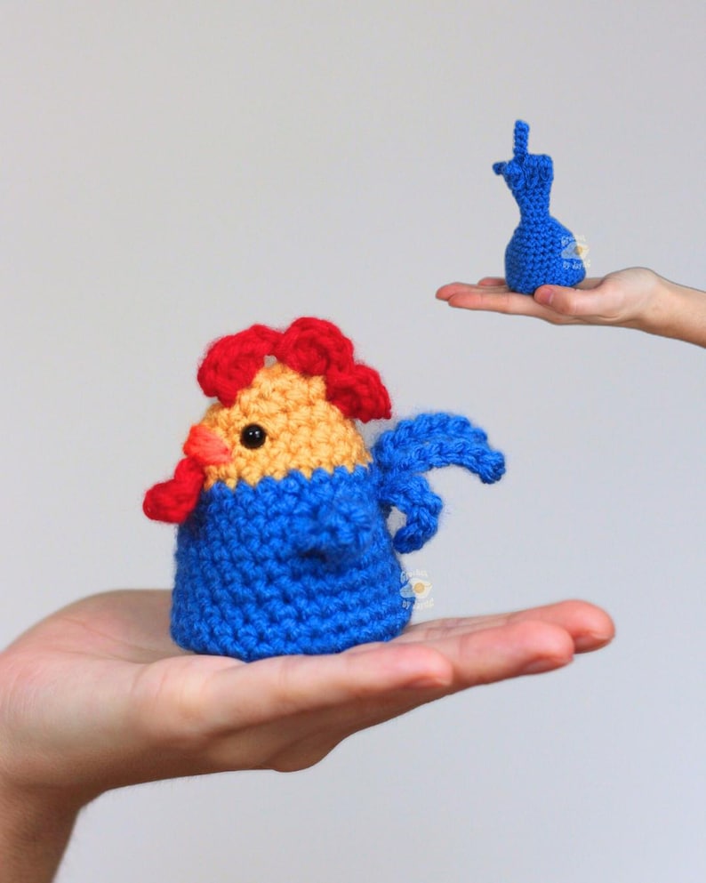 May include: A handmade crochet rooster in blue, yellow, and red, held in a hand. The rooster has a blue body, yellow head, and red comb and wattle. A smaller blue crochet figure is held in another hand.