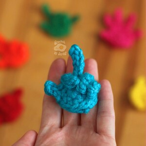 May include: A close-up of a turquoise blue crocheted middle finger, held in a hand. Other crocheted middle fingers in red, green, pink, and yellow are blurred in the background. The text "Crochet by Jayne" and "Etsy Crochet by Jayne" are visible.