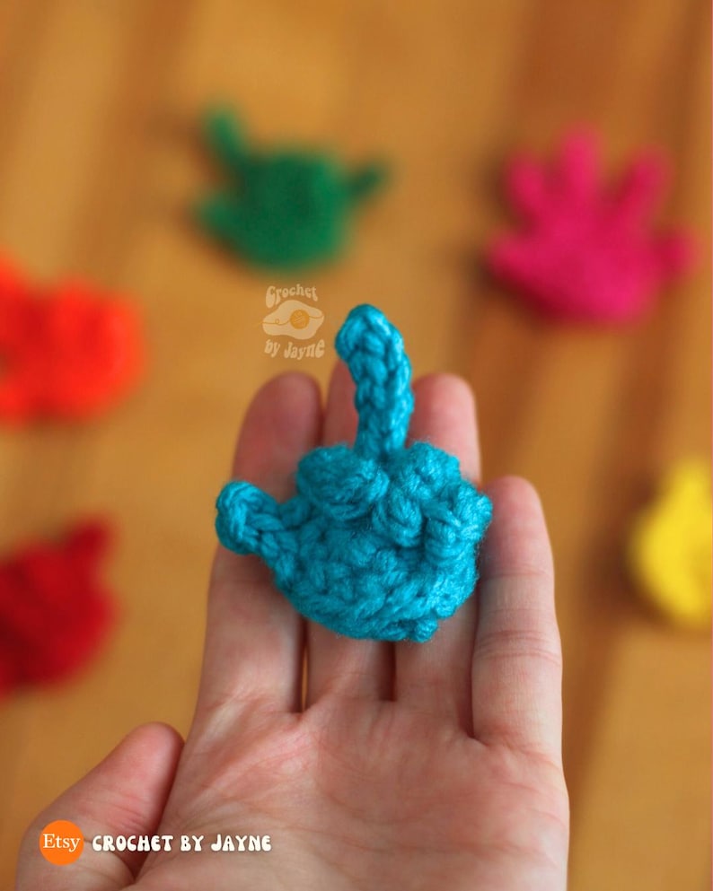May include: A close-up of a turquoise blue crocheted middle finger, held in a hand. Other crocheted middle fingers in red, green, pink, and yellow are blurred in the background. The text "Crochet by Jayne" and "Etsy Crochet by Jayne" are visible.