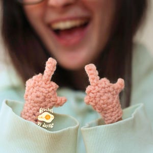 May include: Two peach-colored, crocheted finger puppets, each displaying a raised middle finger. The puppets are emerging from the sleeves of a light green sweatshirt. The text "Crochet by Jayne" is visible on a small tag.
