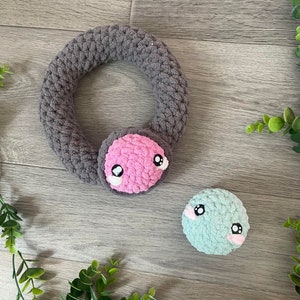 May include: Two crocheted plush toys with embroidered eyes. One is a gray ring with a pink face and the other is a teal ball.