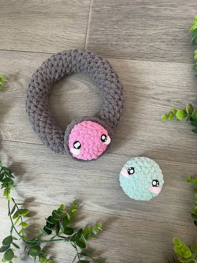 May include: Two crocheted plush toys with embroidered eyes. One is a gray ring with a pink face and the other is a teal ball.