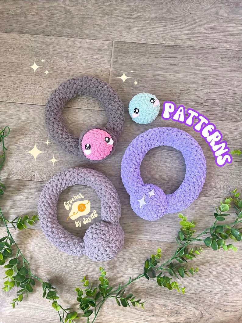 May include: Three crocheted rings with different colored yarn and embellishments. The rings are gray, purple, and light blue. The gray ring has a pink face with eyes and a mouth. The purple ring has two white stars. The light blue ring has a white face with eyes and a mouth. The text "PATTERNS" is written in white letters on a pink background. The text "Crochet by Jayne" is written in white letters on a yellow background.