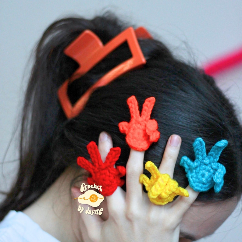 May include: A collection of crochet finger puppets in red, yellow, and blue, shaped like peace signs. The puppets are worn on fingers, with an orange headband in the background. The image includes the text "Crochet by Jayne".