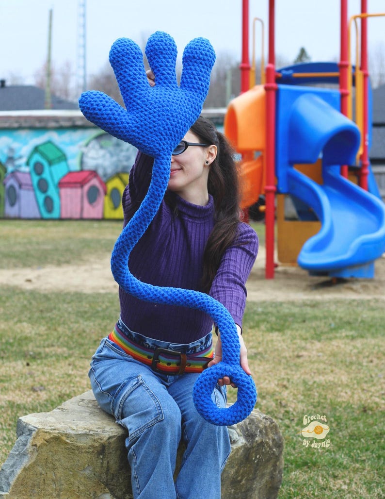 May include: A blue crocheted hand with a long, flexible arm and a ring at the end. The hand is being held by a person sitting on a rock in a playground.