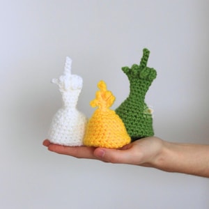 May include: Three crocheted figures in white, yellow, and green, each with a hand gesture. The figures are held in a hand against a plain background. The figures are handmade and have a unique texture.