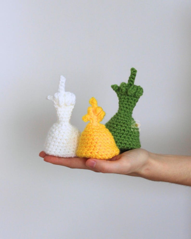 May include: Three crocheted figures in white, yellow, and green, each with a hand gesture. The figures are held in a hand against a plain background. The figures are handmade and have a unique texture.