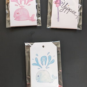 May include: Three white gift tags with blue and pink watercolor illustrations of whales and the word "Yay" in blue and pink. The tags are packaged in clear plastic bags.