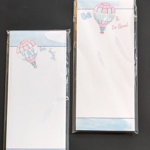 May include: Two white notepads with blue and pink watercolor designs of hot air balloons. One notepad says "Be Happy & Do Good" and the other says "Yay for Today!"