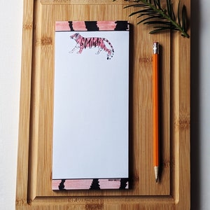 May include: A notepad with a pink and black tiger design and the words "Go Tigers" on the front. The notepad is on a wooden cutting board with a pencil next to it.