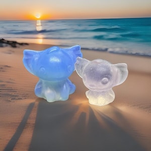Może przedstawiać: Dwie przezroczyste, kreskówkowe figurki, jedna niebieska i jedna lawendowa, stoją na piaszczystej plaży z oceanem w tle. Figurki mają duże uszy i są podświetlane od wewnątrz, rzucając cienie. Scena rozgrywa się o zachodzie słońca.