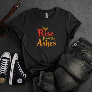 May include: A dark grey t-shirt with a graphic design of a phoenix rising from the ashes. The text "Rise from the Ashes" is printed in red and yellow.