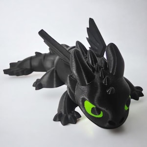 Puede incluir: Figura de dragón negro con ojos verde brillante. El dragón tiene alas detalladas, rasgos puntiagudos y una larga cola. La figura está tumbada sobre una superficie blanca. Este dragón impreso en 3D es un objeto de colección.