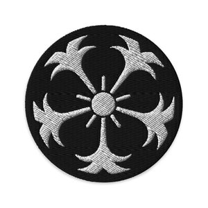 May include: A black and white embroidered patch with a stylized sun and four radiating arms. The patch is circular and has a black background with white embroidery.