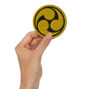 Peut inclure: Un écusson brodé jaune avec un symbole noir en trois parties, ressemblant à une fleur stylisée ou un motif géométrique.