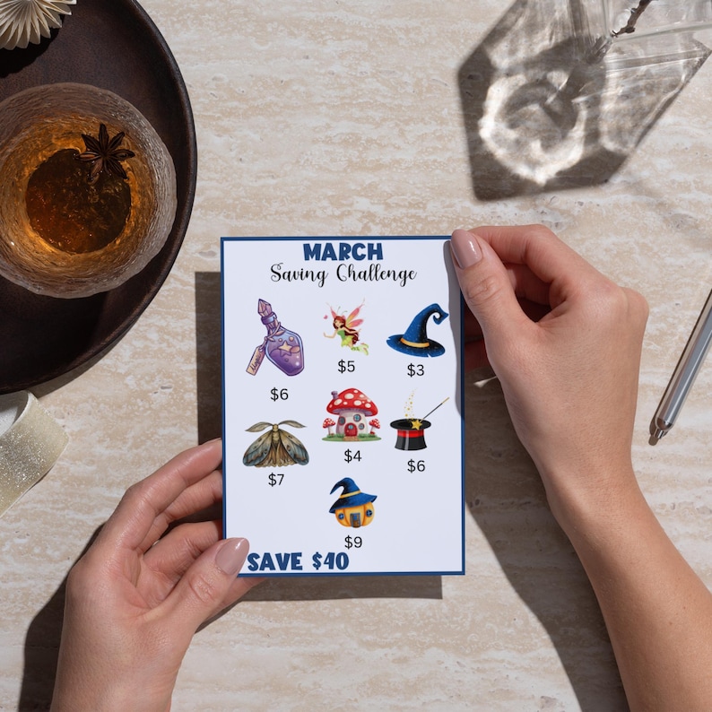 A person's hands are holding a card with various saving challenge icons, including a mushroom, fairy, witch's hat, and other whimsical items, along with the text "MARCH Saving Challenge" and the amount "$9".