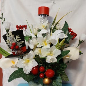 May include: A white flower arrangement in a silver pot with a black top hat and red berries. The arrangement includes white calla lilies and white orchids.