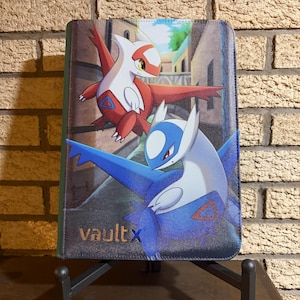 Pode incluir: Um fichário retangular para cartas colecionáveis com o logotipo Vault X e uma ilustração gráfica dos personagens Latias vermelho e Latios azul contra um fundo de uma rua de pedra com um canal.