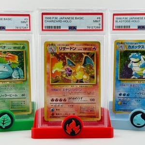 Może przedstawiać: Trzy karty kolekcjonerskie Pokemon w ochronnych etui. Karty to japońskie wersje Venusaura, Charizarda i Blastoise'a. Każda karta jest kartą holograficzną z oceną stanu idealnego.