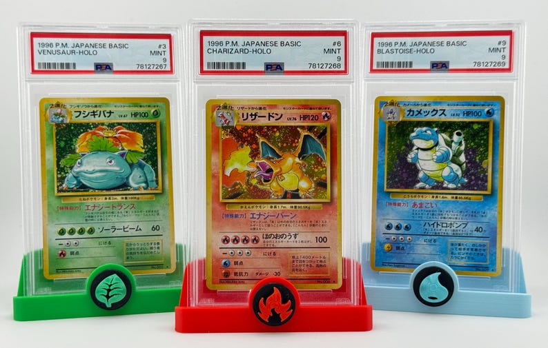 Może przedstawiać: Trzy karty kolekcjonerskie Pokemon w ochronnych etui. Karty to japońskie wersje Venusaura, Charizarda i Blastoise'a. Każda karta jest kartą holograficzną z oceną stanu idealnego.