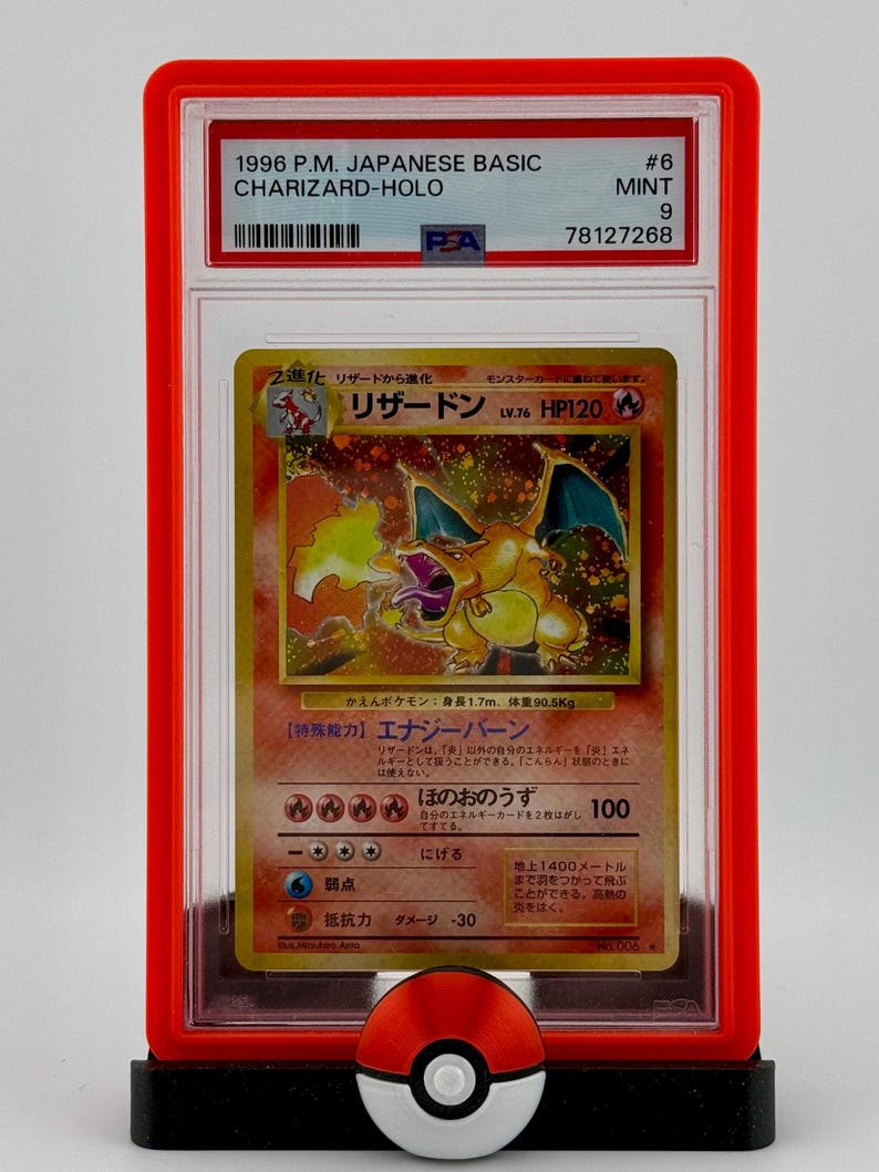 Może przedstawiać: Zgradeda karta kolekcjonerska Pokemon w ochronnym etui. Karta to japoński Charizard-Holo z zestawu 1996 P.M. Basic. Karta została oceniona na 9 przez PSA, co oznacza, że ​​jest w doskonałym stanie.