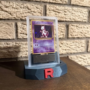 May include: A clear plastic display case with a gold trim, showcasing a Japanese Pokemon card featuring a purple and white Mewtwo. The case is sitting on a gray 3D printed stand with a red "R" on the front.