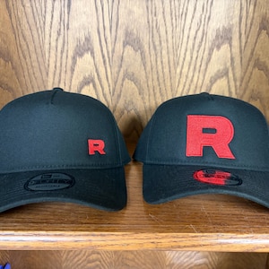 Puede incluir: Dos gorras de béisbol negras con coronas estructuradas y viseras curvas. La gorra de la izquierda presenta una pequeña letra R roja bordada, mientras que la de la derecha muestra una letra R roja grande bordada. Ambas tienen pegatinas.