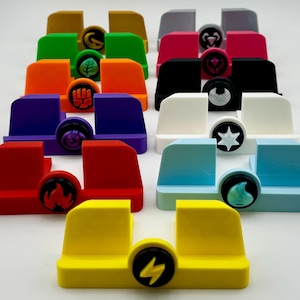 Może przedstawiać: Zestaw 12 kolorowych stojak&oacute;w na telefon z r&oacute;żnymi symbolami z przodu. Stojaki są wykonane z tworzywa sztucznego i są zaprojektowane do trzymania telefonu w pozycji pionowej.