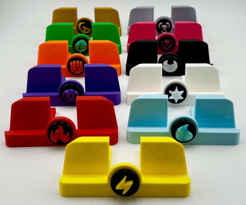 Może przedstawiać: Zestaw 12 kolorowych stojak&oacute;w na telefon z r&oacute;żnymi symbolami z przodu. Stojaki są wykonane z tworzywa sztucznego i są zaprojektowane do trzymania telefonu w pozycji pionowej.